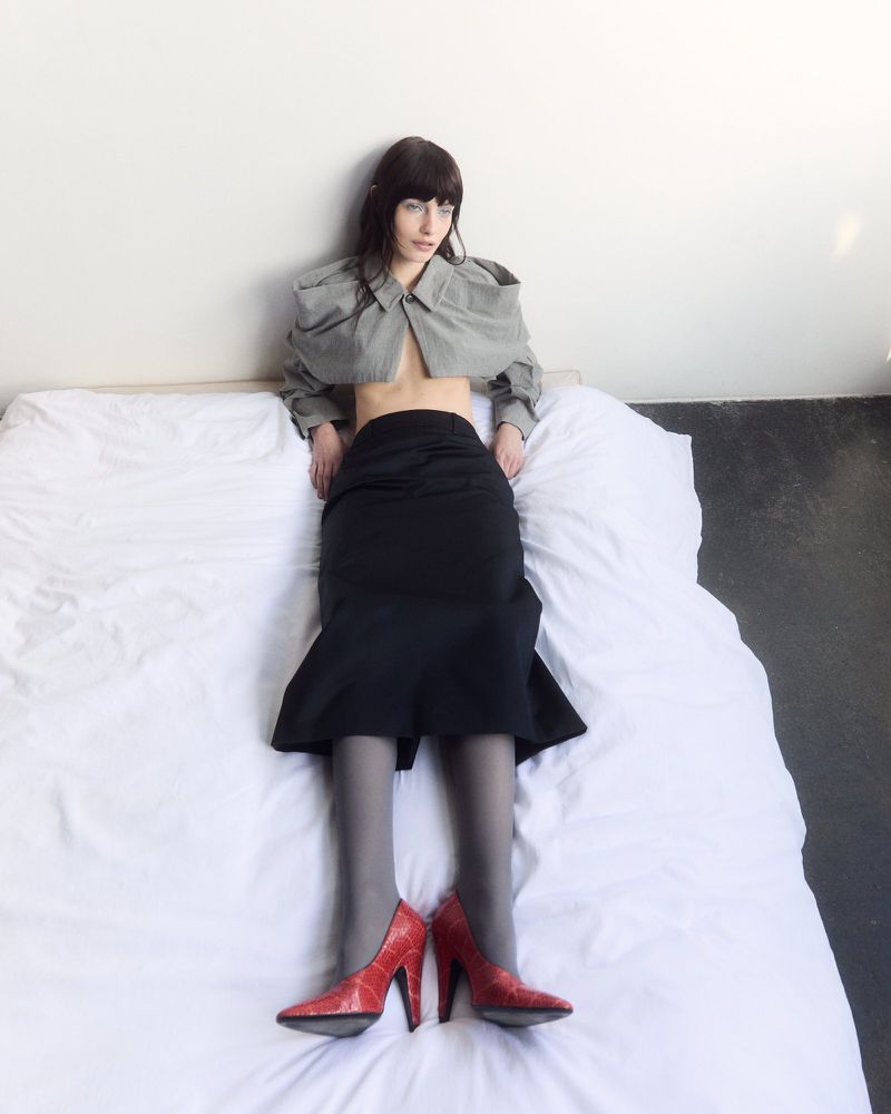 Clara Denison in a cropped grey jacket, black midi skirt, and red heels reclining on a white sheet against a neutral studio wall.