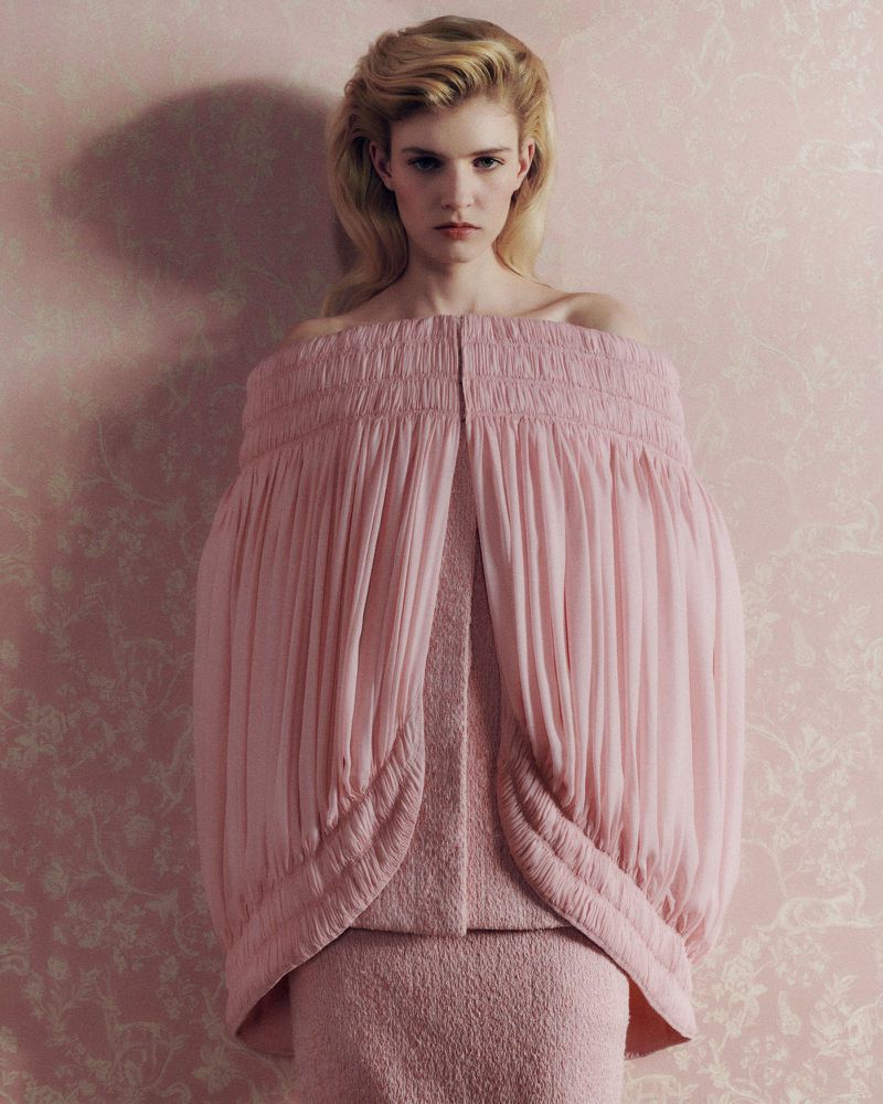Model Camille Chifflot in a voluminous off-the-shoulder pink pleated cape garment against floral pink wallpaper.