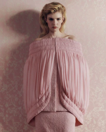 Model Camille Chifflot in a voluminous off-the-shoulder pink pleated cape garment against floral pink wallpaper.