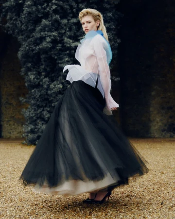 Model in voluminous black tulle skirt and sheer layered blouse posing outdoors against dark foliage and gravel ground.