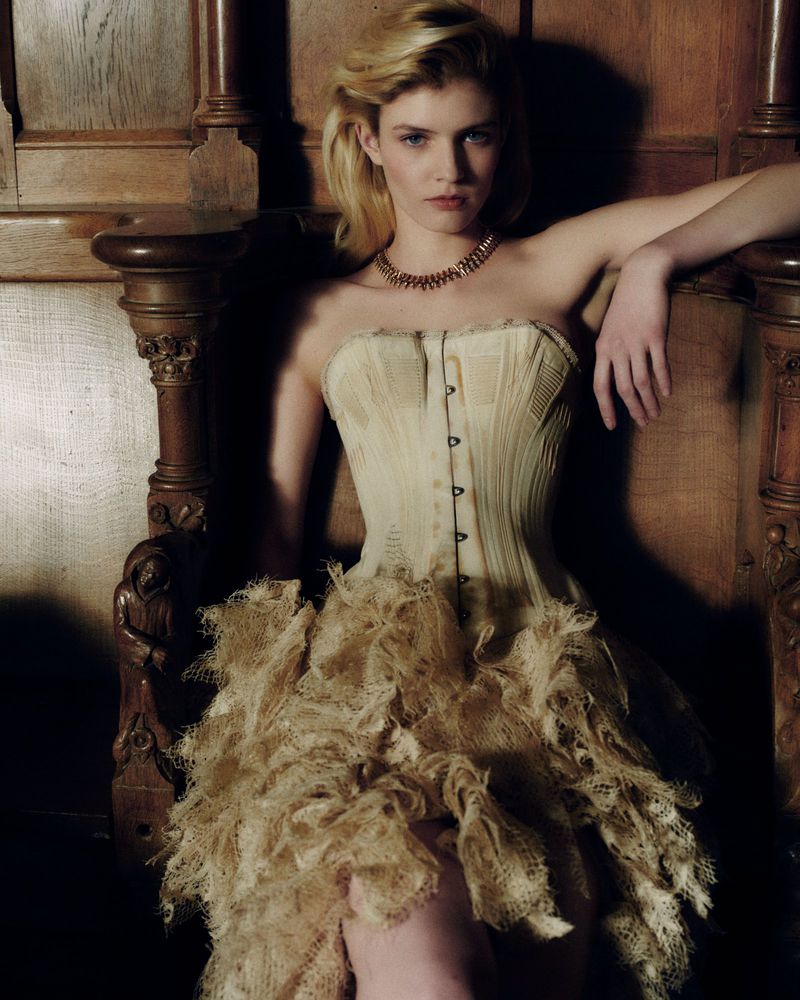 Model Camille Chifflot wearing a structured cream corset dress and gold necklace while seated in a carved wooden high-backed chair.
