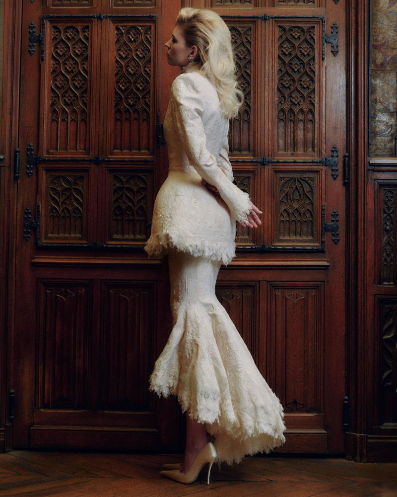 Model in textured white dress standing in profile before ornate carved wooden doors for Telegraph Luxury
