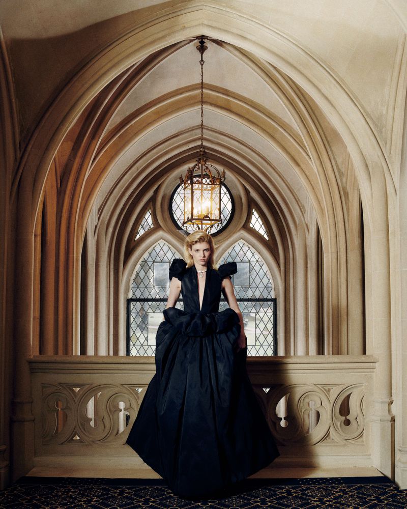 Model Camille Chifflot in a voluminous black gown standing under gothic stone arches with a hanging lantern chandelier.