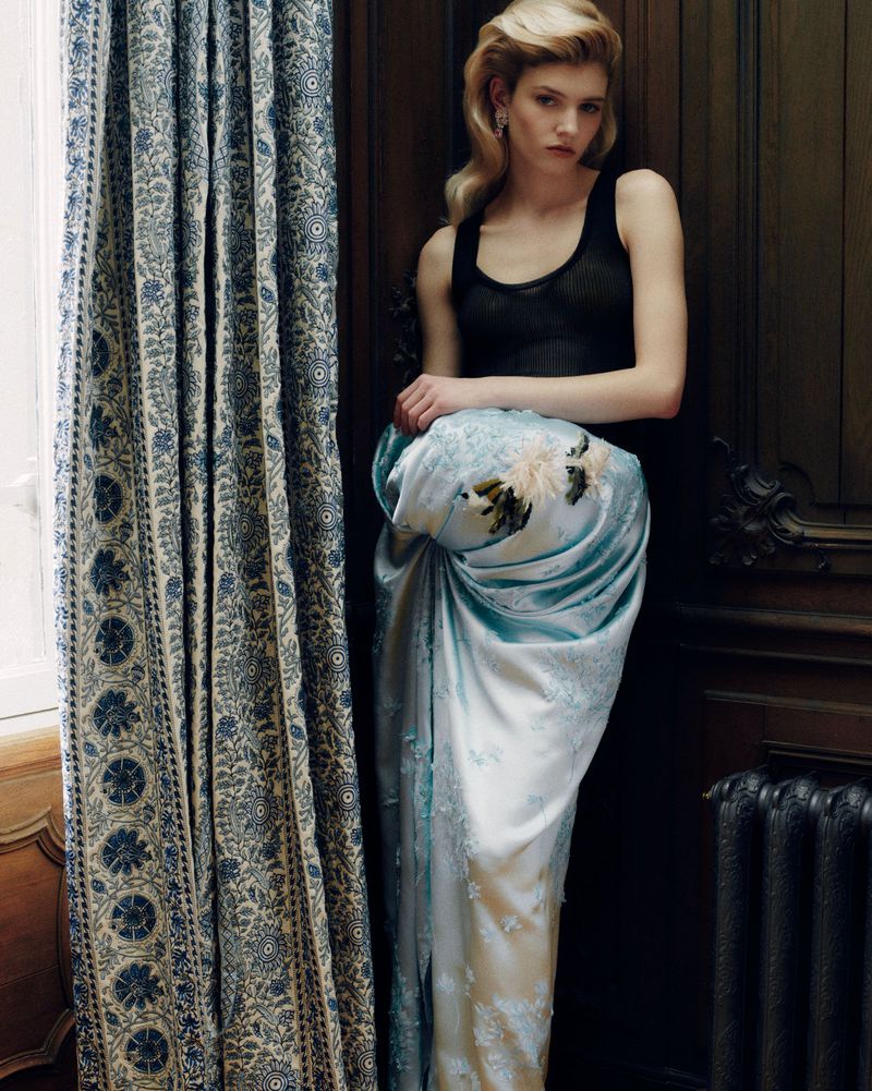 Model leaning against a dark wood wall next to blue patterned curtains, wearing a black top and a draped light blue skirt.