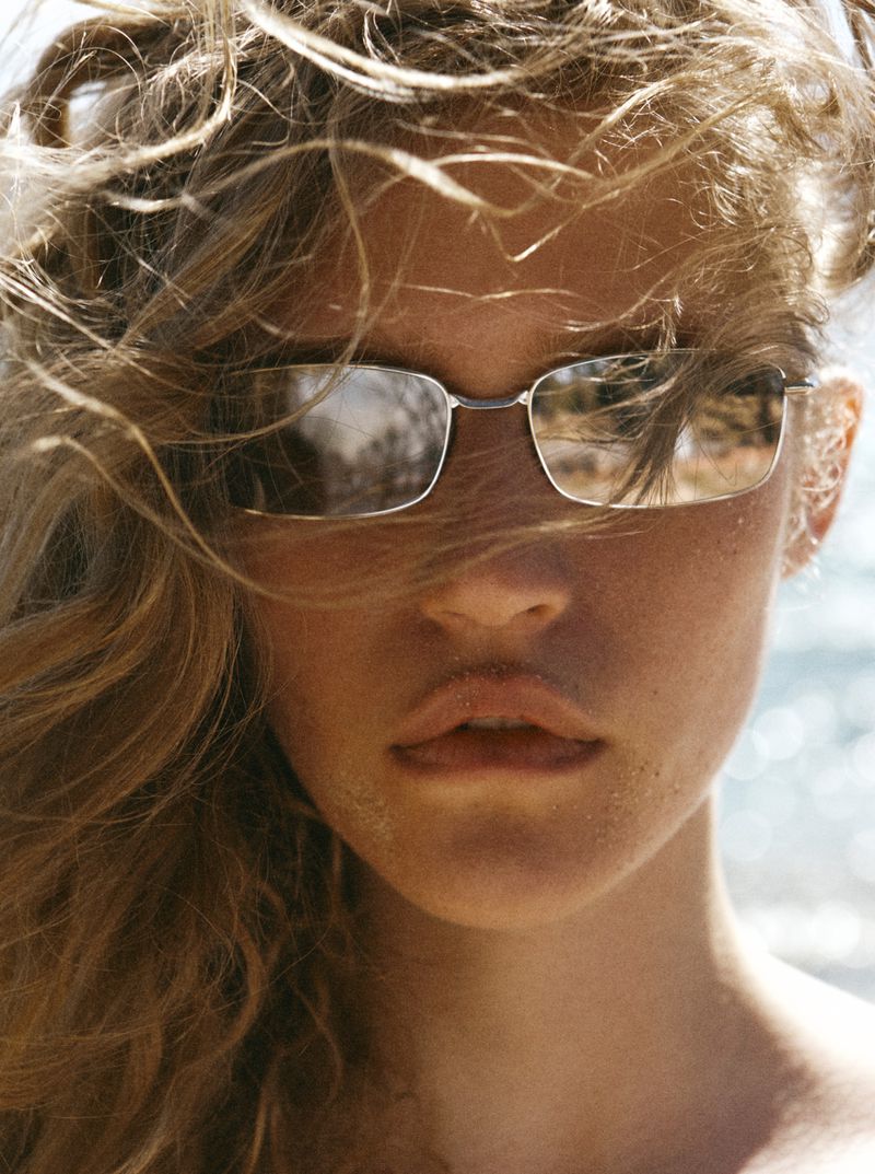 Close-up portrait of model Mariam de Vinzelle wearing rectangular wire-frame sunglasses with windblown hair outdoors.