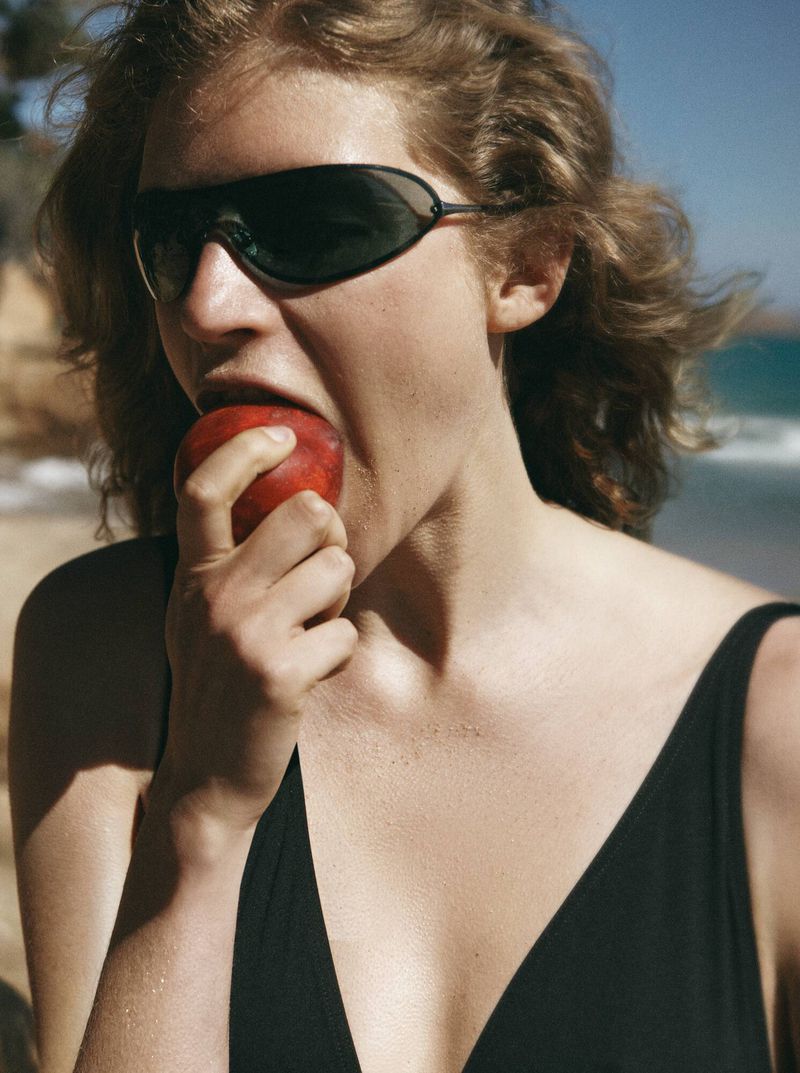Model Mariam de Vinzelle wearing sporty black sunglasses and a black swimsuit while biting into a peach at the beach.