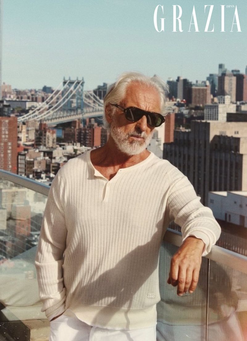 Gil Ros wearing dark sunglasses and a cream ribbed sweater leaning on a balcony with a city skyline background.