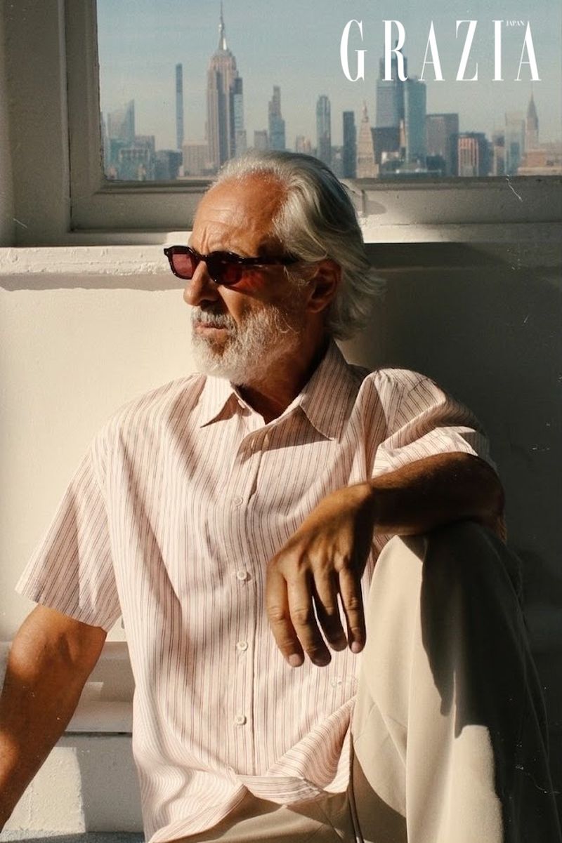 Model Gil Ros with grey beard wearing sunglasses and a striped short-sleeve shirt seated by a window with a city skyline view.