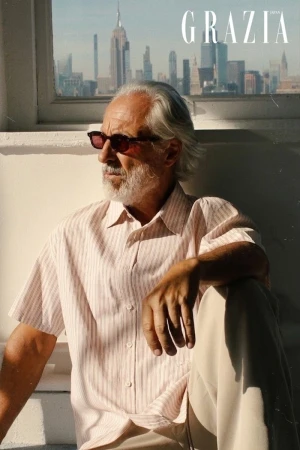 Model Gil Ros with grey beard wearing sunglasses and a striped short-sleeve shirt seated by a window with a city skyline view.