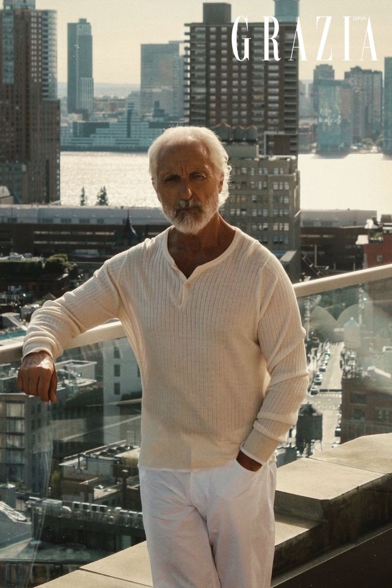 Gil Ros standing on an urban rooftop balcony wearing a cream long-sleeve knit shirt and white trousers.