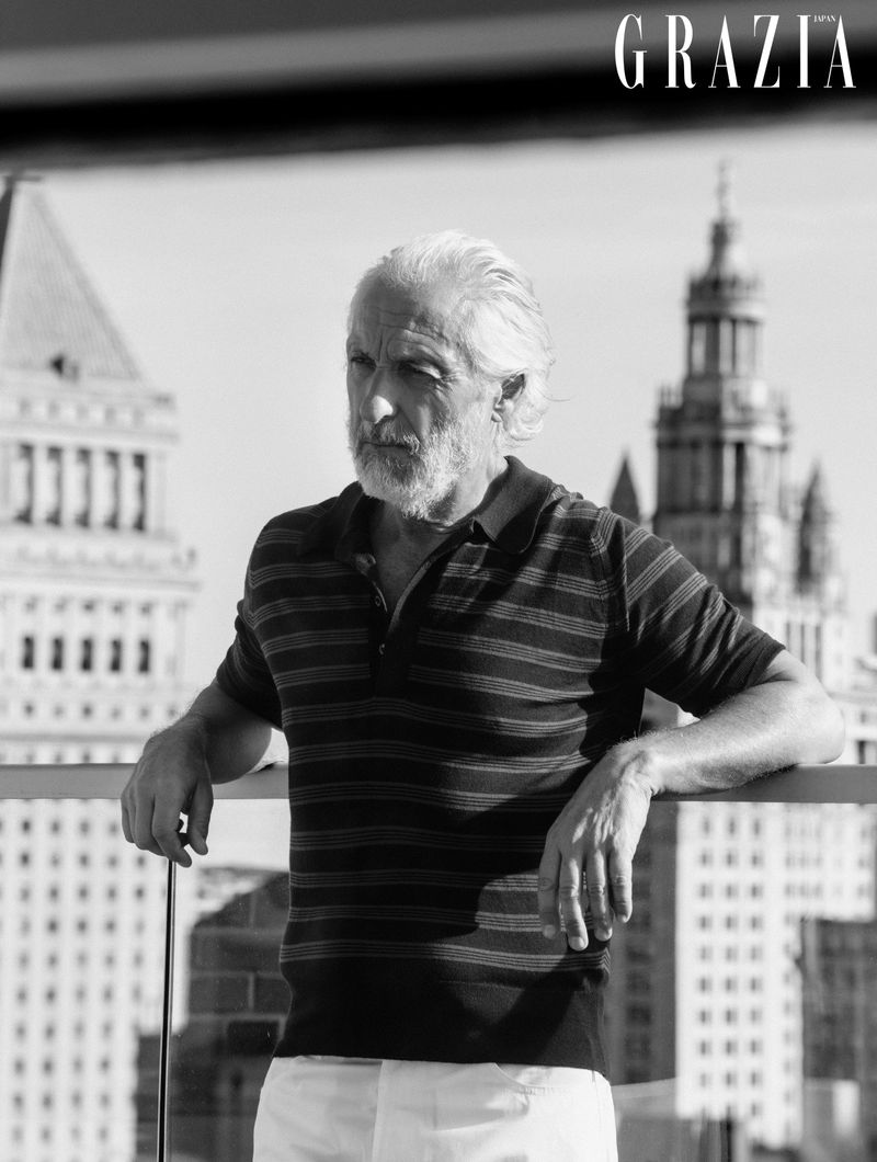 Mature man with white hair and beard wearing a striped polo shirt, leaning on a rooftop railing with city buildings in background.
