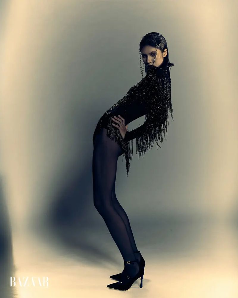 Model posing in a black fringe bodysuit, dark tights, and pointed high heels against a neutral studio backdrop.