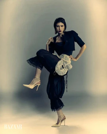 Fashion model in a black cropped jacket, dark bloomers, and nude slingback stiletto heels posing with one leg raised in a studio.