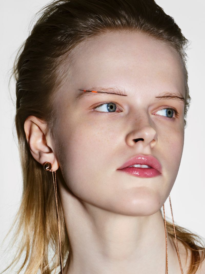 Close-up portrait of a fair-skinned woman with long blonde hair, wearing a pair of gold earrings against a white background.