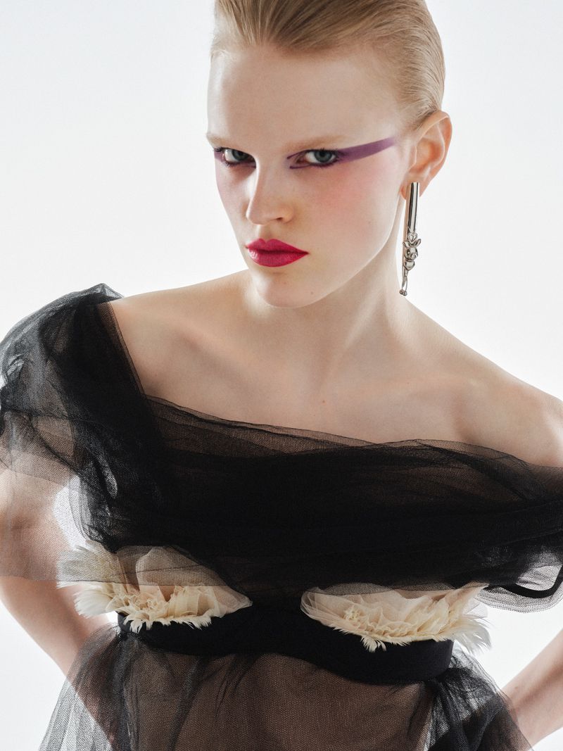 Portrait of fair-skinned model wearing avant-garde makeup and a black tulle dress against a bright white backdrop in a studio setting.