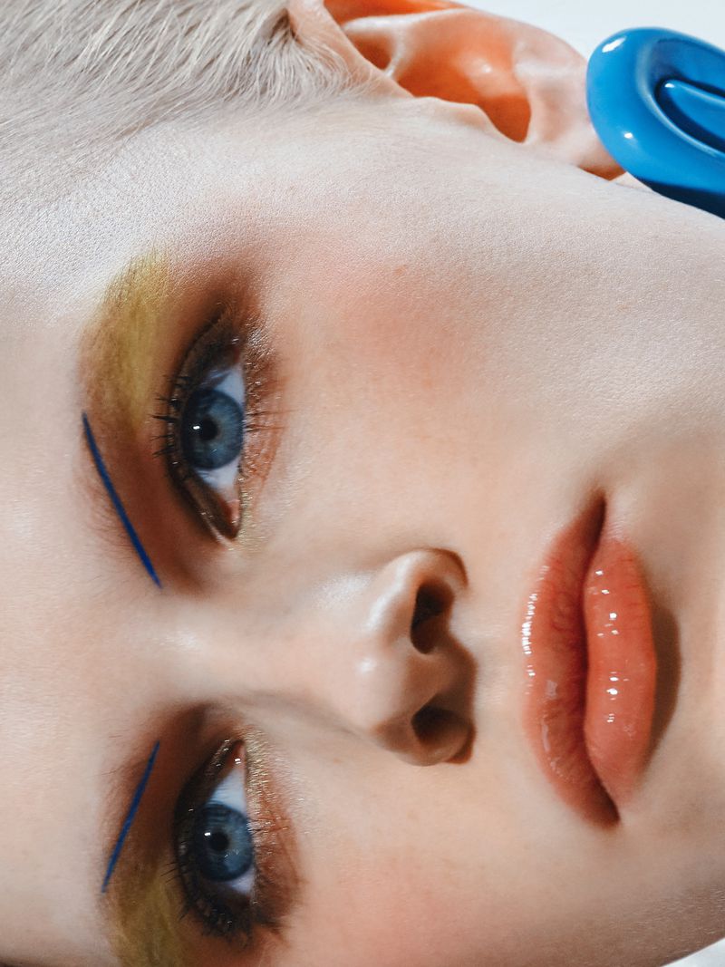 Close-up portrait of a young adult woman with blue eyes, light hair, and makeup, wearing a blue accessory, looking at the camera.