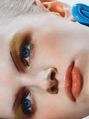 Close-up portrait of a young adult woman with blue eyes, light hair, and makeup, wearing a blue accessory, looking at the camera.