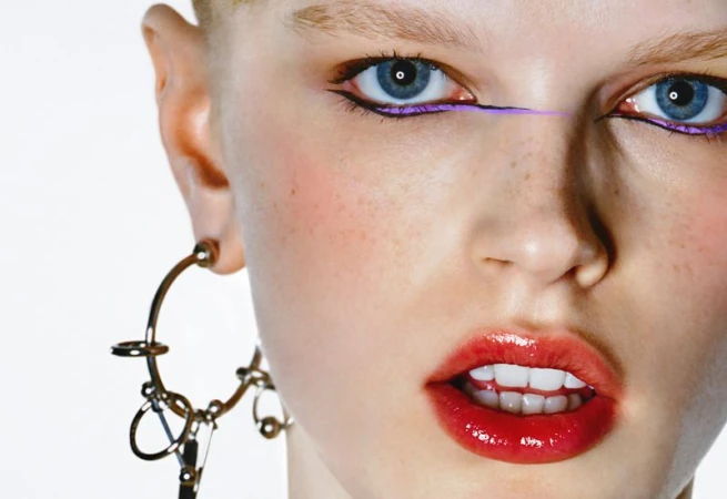 Close-up portrait of a fair-skinned woman with blue eyes, wearing bold makeup with purple eyeliner and glossy red lipstick against a white background.