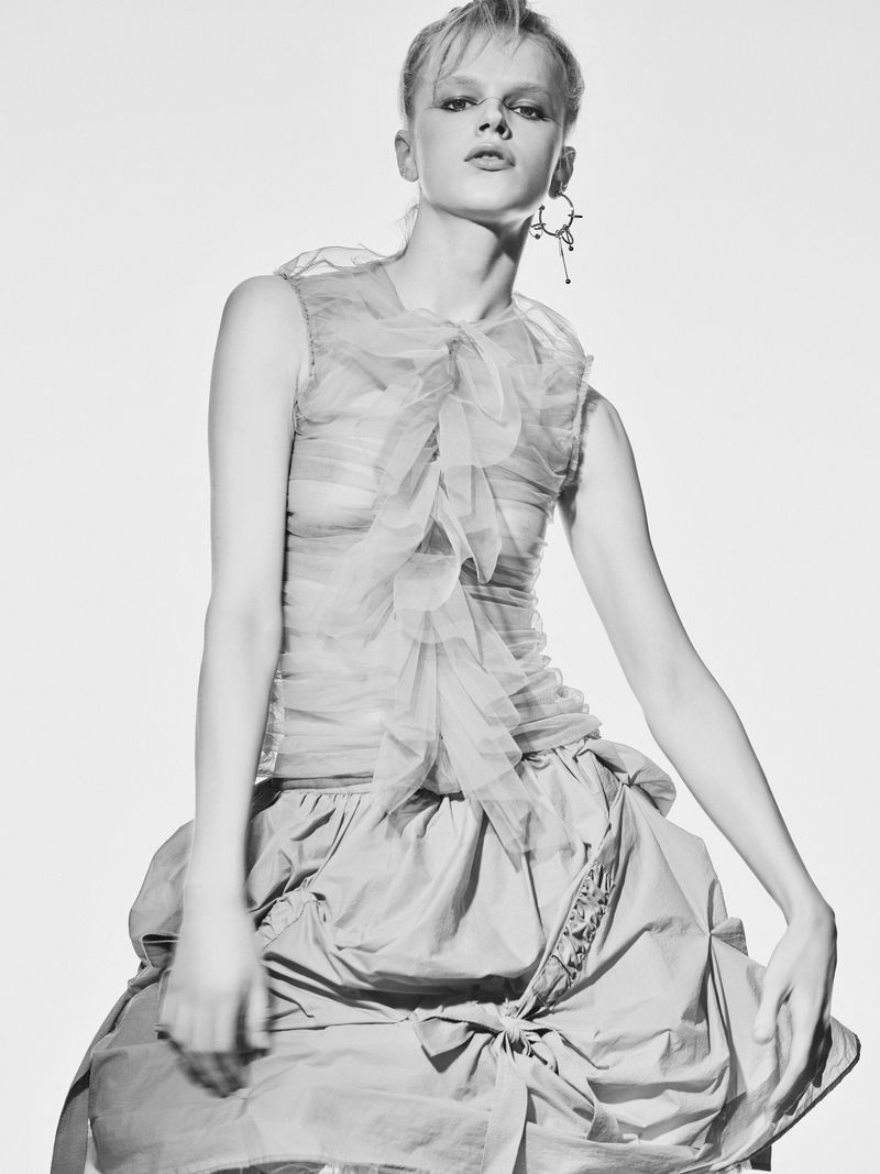 Black and white fashion shot of a model wearing a layered top with ruffles and a full skirt, posing in a brightly lit photo studio.