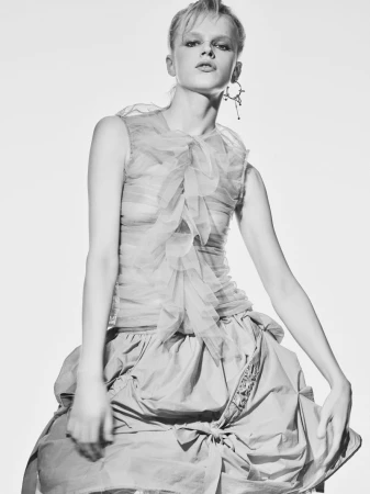 Black and white fashion shot of a model wearing a layered top with ruffles and a full skirt, posing in a brightly lit photo studio.