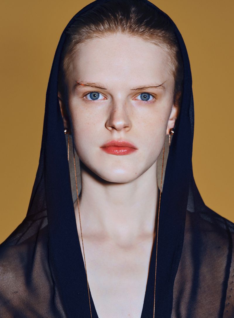 Close-up of a blue-eyed woman with sheer black hooded top, long gold earrings, and coral lipstick against yellow-gold backdrop.