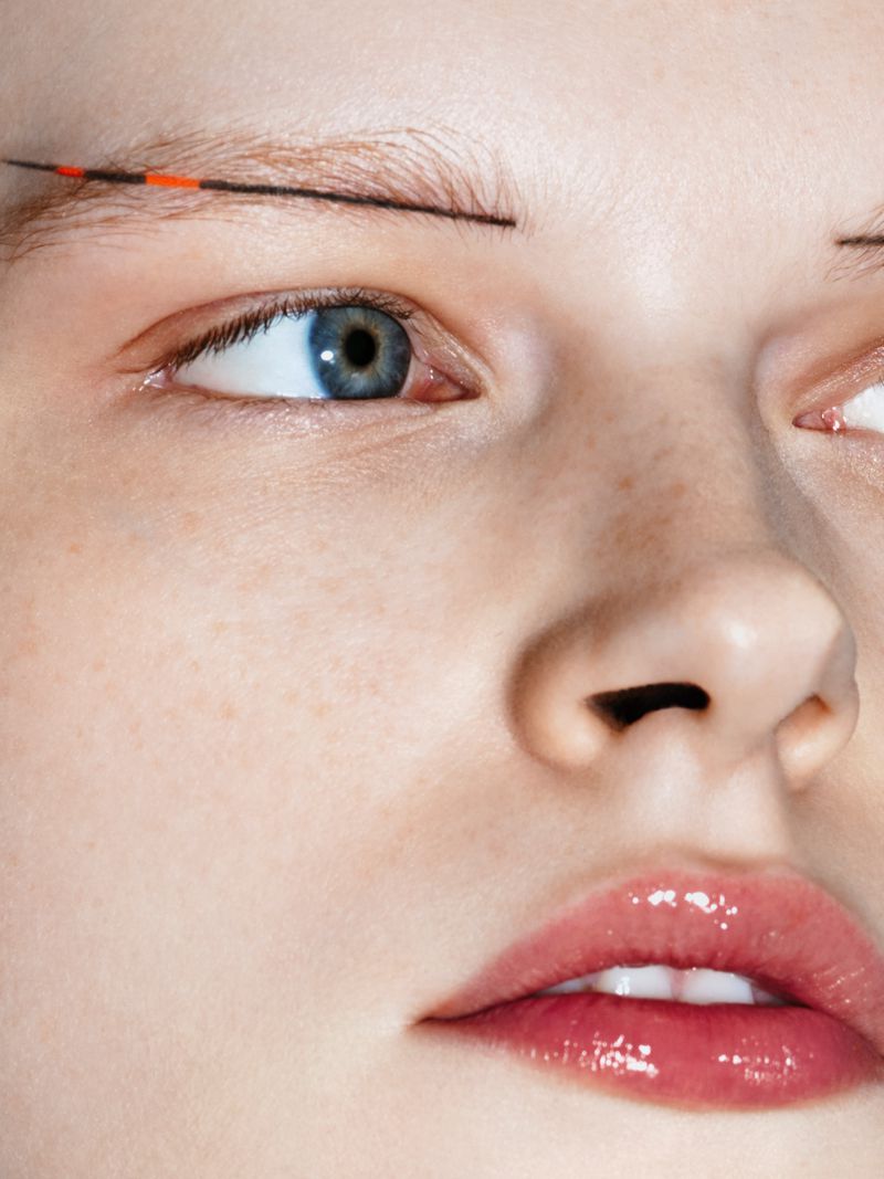 Close-up studio shot of a woman's face with minimalist graphic makeup using thin, black lines and an orange accent on her eyebrow.