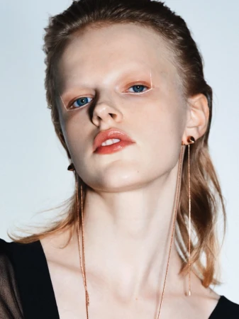 Close-up studio shot of a young fair-skinned woman with light blonde hair, glossy lips, dramatic eye makeup, and wearing gold dangling earrings.