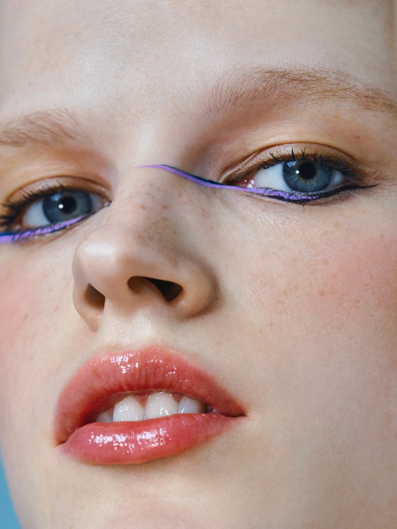 Close-up shot of a fair-skinned young woman with blue eyes and glossy lips, wearing purple eyeliner on a light blue background.