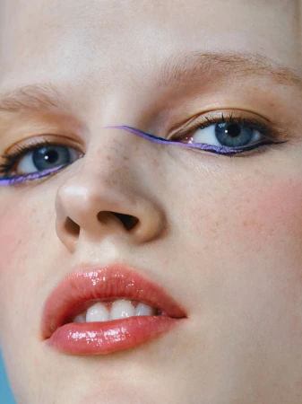 Close-up shot of a fair-skinned young woman with blue eyes and glossy lips, wearing purple eyeliner on a light blue background.