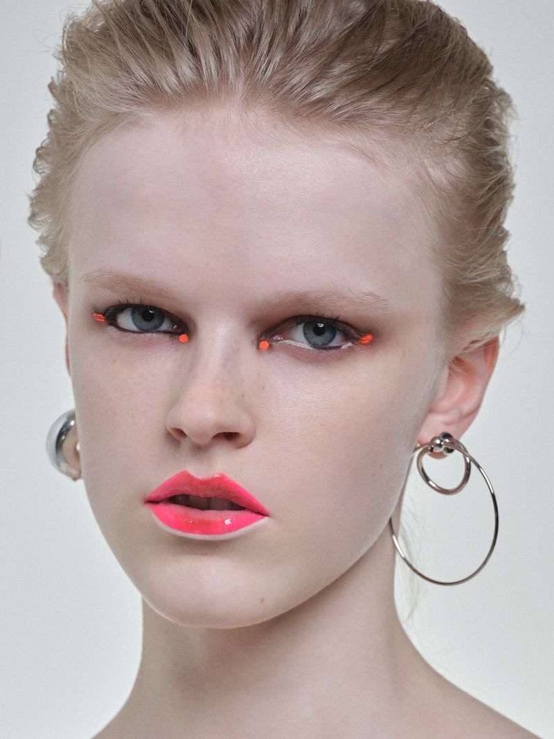 Close-up portrait of a fair-skinned woman with neon pink lipstick, silver hoop earrings, and creative orange makeup in a light studio.
