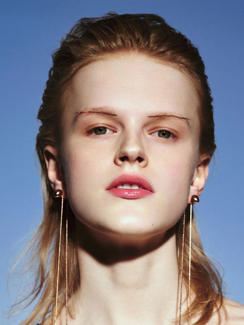 Close-up portrait of a young fair-skinned woman with light brown hair, wearing earrings against a clear blue sky backdrop.