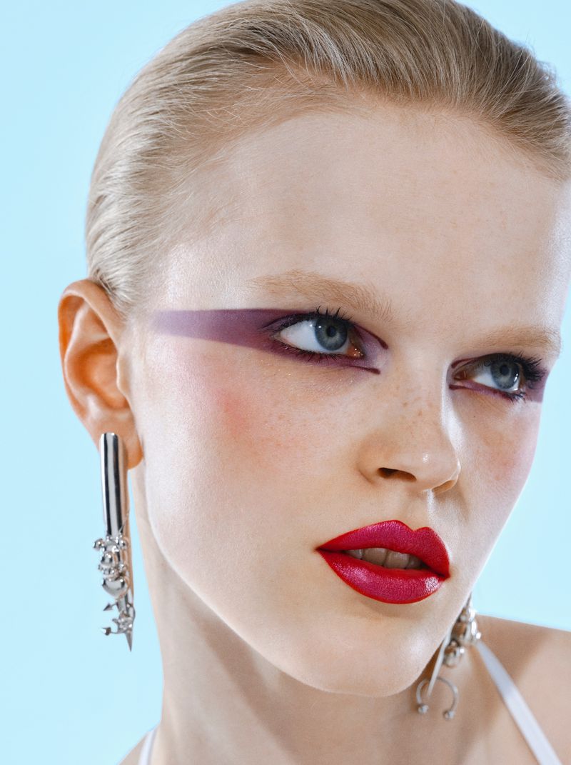 Close-up of a fair-skinned woman with sleek blonde hair, striking magenta eyeshadow, and bold red lipstick, wearing silver earrings against light blue.