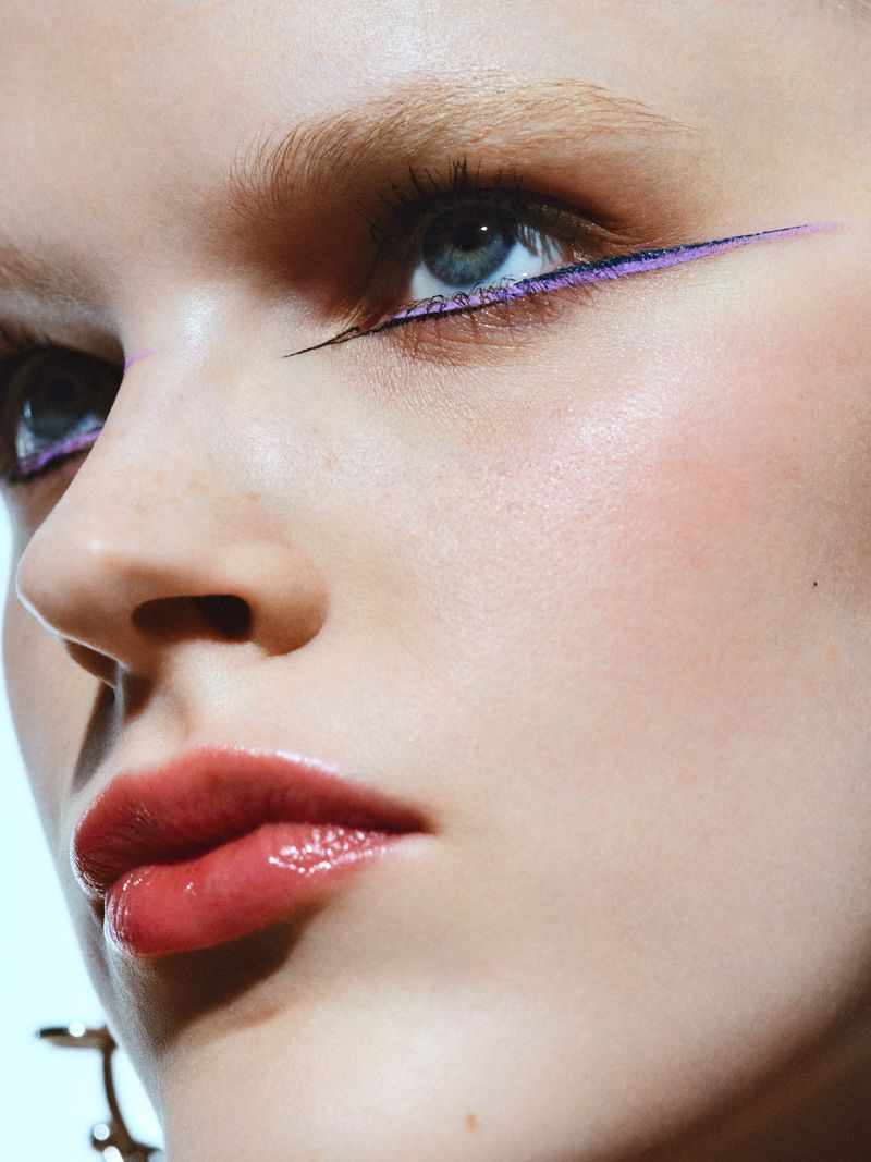 Close-up portrait of a young adult woman with blue eyes, wearing purple eyeliner and lipstick against a light background in a beauty shot.
