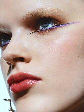 Close-up portrait of a young adult woman with blue eyes, wearing purple eyeliner and lipstick against a light background in a beauty shot.