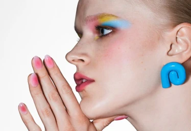 Close-up profile of a model with colorful eye makeup, pink gradient nails, and a bold blue earring against a white background.