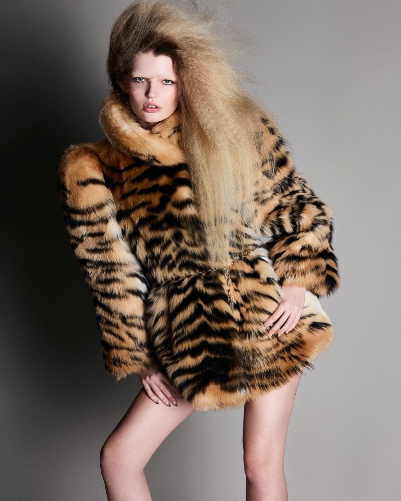 Model with voluminous crimped blonde hair wearing a tiger stripe faux fur coat against a grey background.