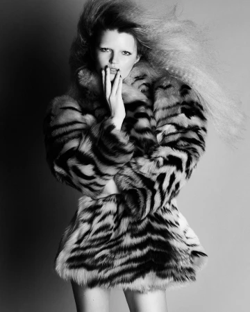 Black and white fashion portrait of a woman with voluminous crimped hair wearing a striped fur coat against a studio backdrop.