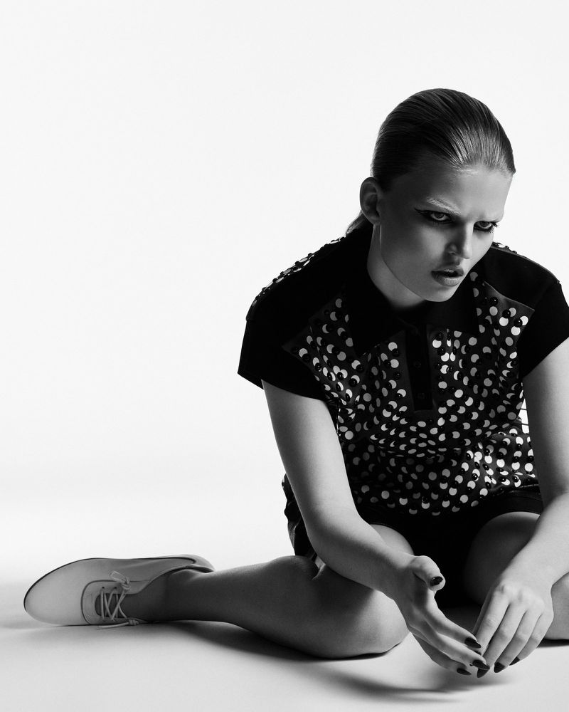 Model wearing a patterned collared top and lace-up shoe sitting on a studio floor in black and white