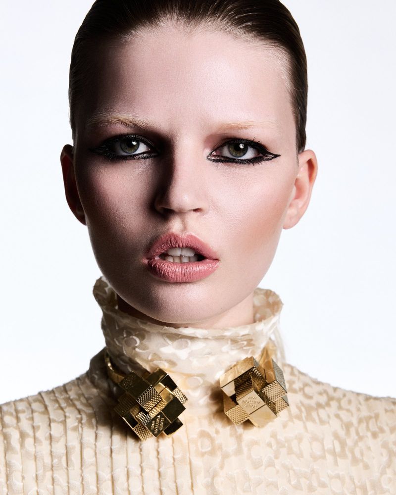 Close-up beauty portrait of a woman with heavy black graphic eyeliner and slicked hair wearing a cream collar with gold geometric jewelry.