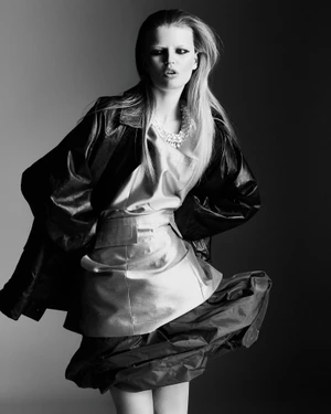 Fashion model wearing an oversized leather jacket and metallic dress with statement necklace against a studio gradient backdrop.
