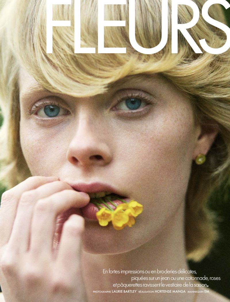 Close-up portrait of a blonde model with blue eyes biting small yellow flowers between her lips.