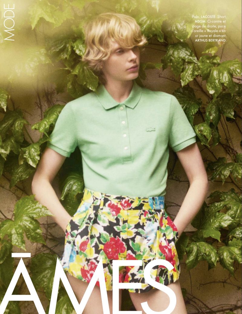 Model Em Verleun in a mint green polo shirt and colorful floral shorts stands with hands in pockets against an ivy-covered wall.