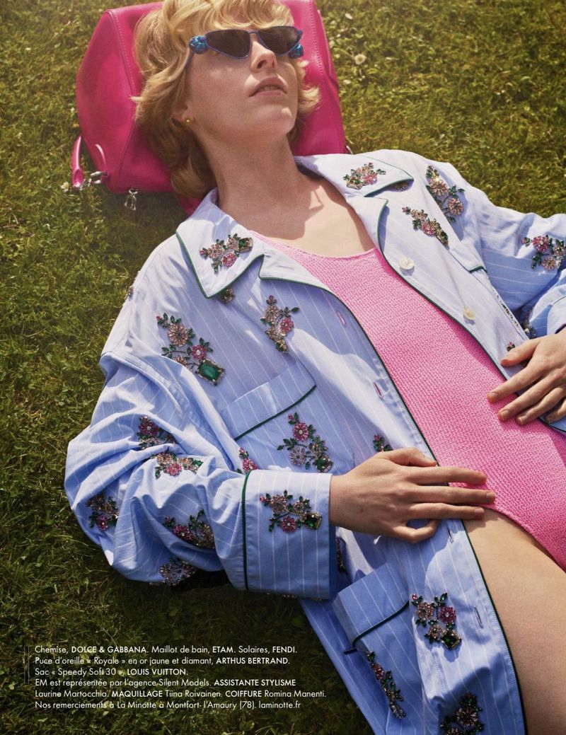 Model Em Verleun lies on green grass wearing an embellished light blue striped shirt, pink swimsuit, and sunglasses against a pink bag.