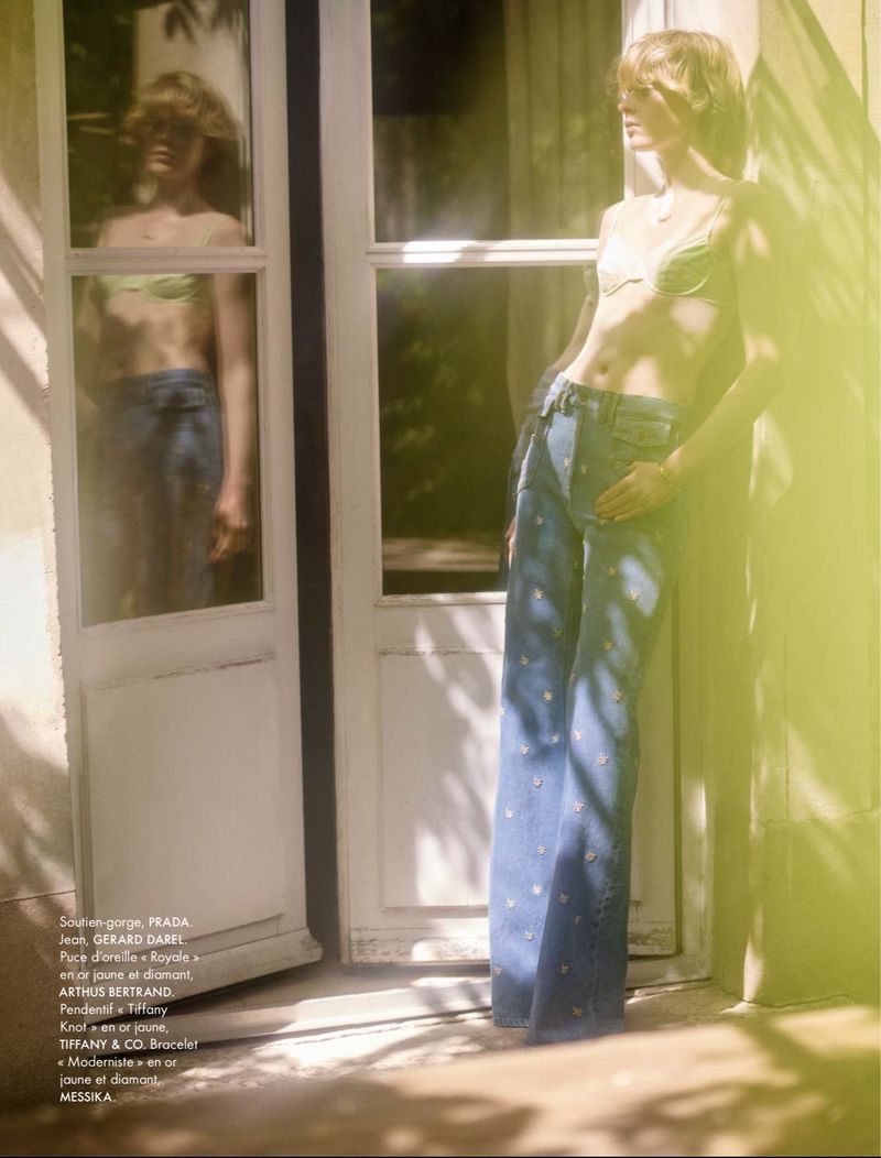 Model Em Verleun posing beside glass French doors wearing a light green bralette and flared blue denim jeans for Elle France.