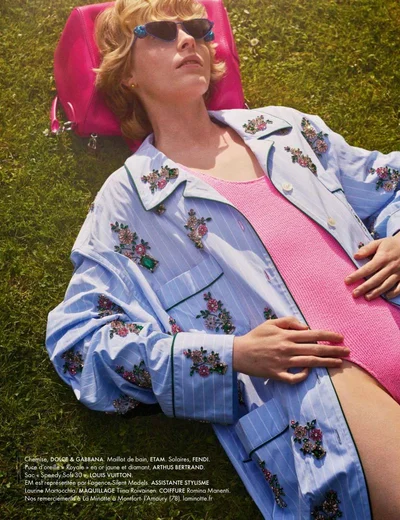 Model Em Verleun lies on green grass wearing an embellished light blue striped shirt, pink swimsuit, and sunglasses against a pink bag.