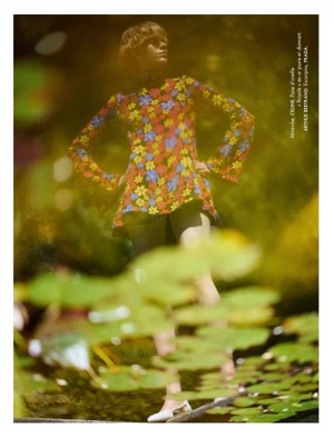 Model Em Verleun wearing a vibrant floral long-sleeve mini dress and white flats beside a pond filled with water lily pads.