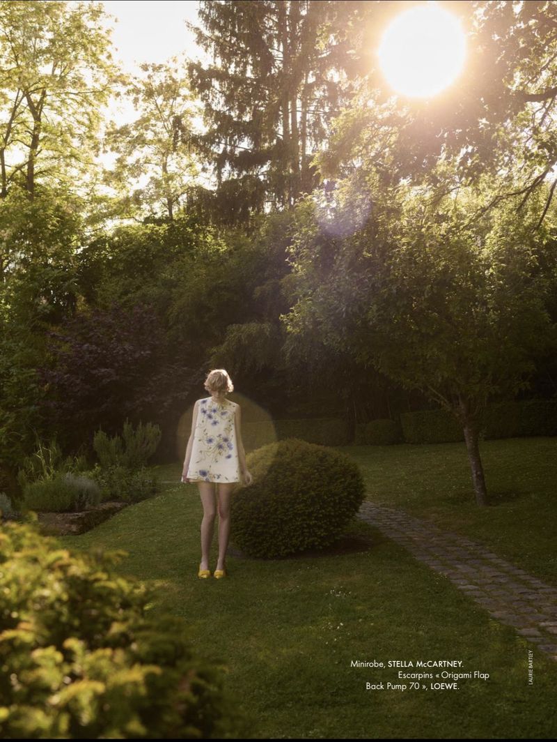 Em Verleun standing in a sunlit garden wearing a sleeveless white floral mini dress and yellow heels, viewed from behind.