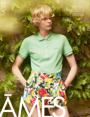 Model Em Verleun in a mint green polo shirt and colorful floral shorts stands with hands in pockets against an ivy-covered wall.
