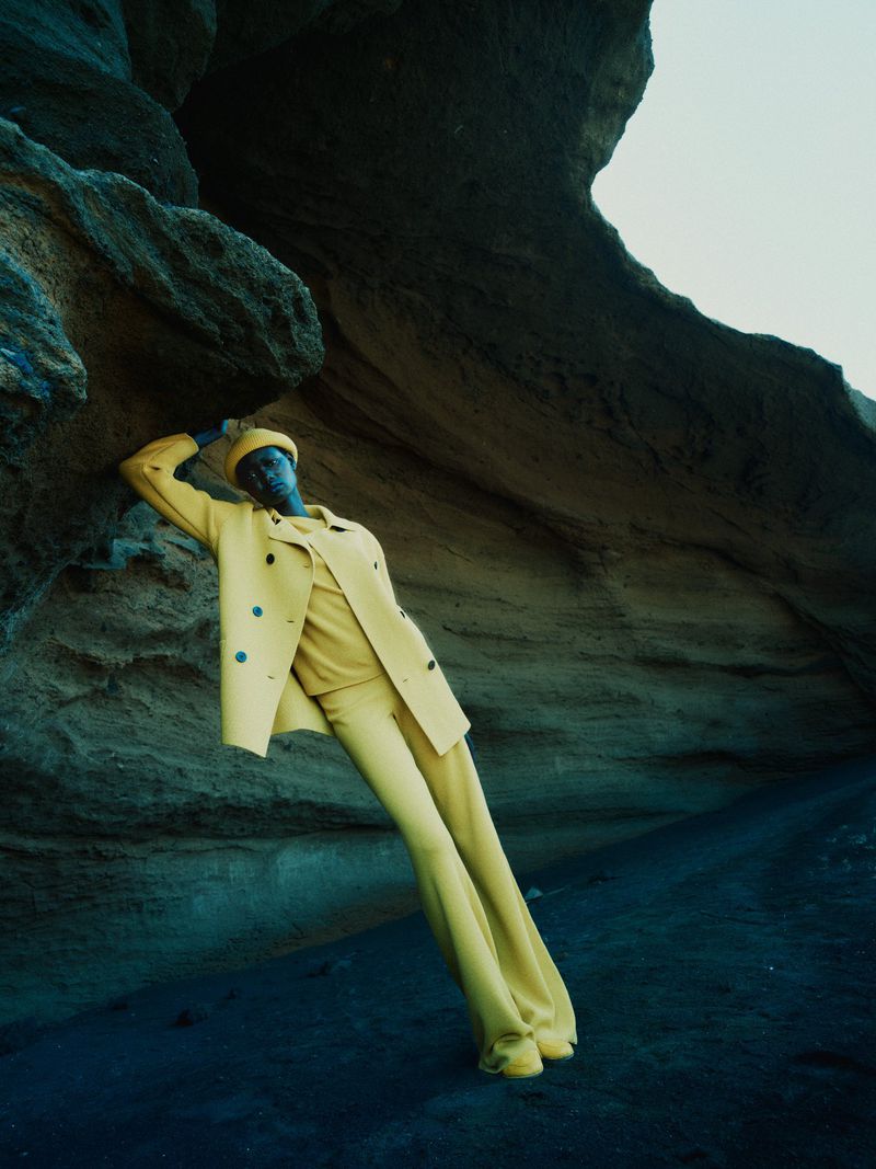 Model Ateta Jok poses leaning against a dark rock wall wearing a vibrant yellow suit, coat, and matching beret.