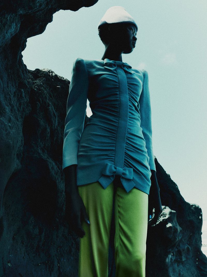 Model Ateta Jok in a light blue ruched dress, lime green trousers, and a white hat framed by dark rocks against a pale sky.